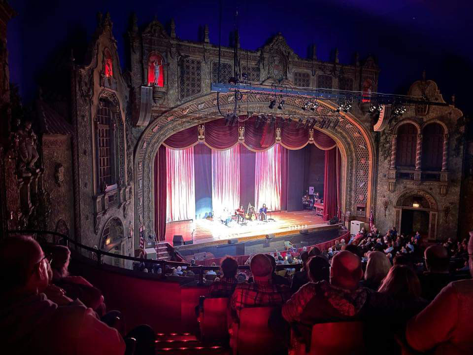 An audience in seats in a large theater viewing a lit stage.