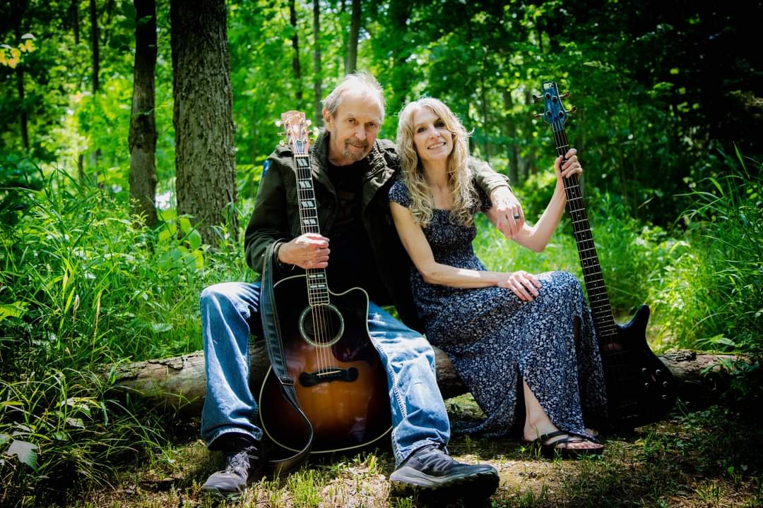 man and woman each holding a guitar while seated on a log in the woods