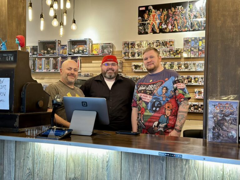 three men featured behind the counter in front of comic books
