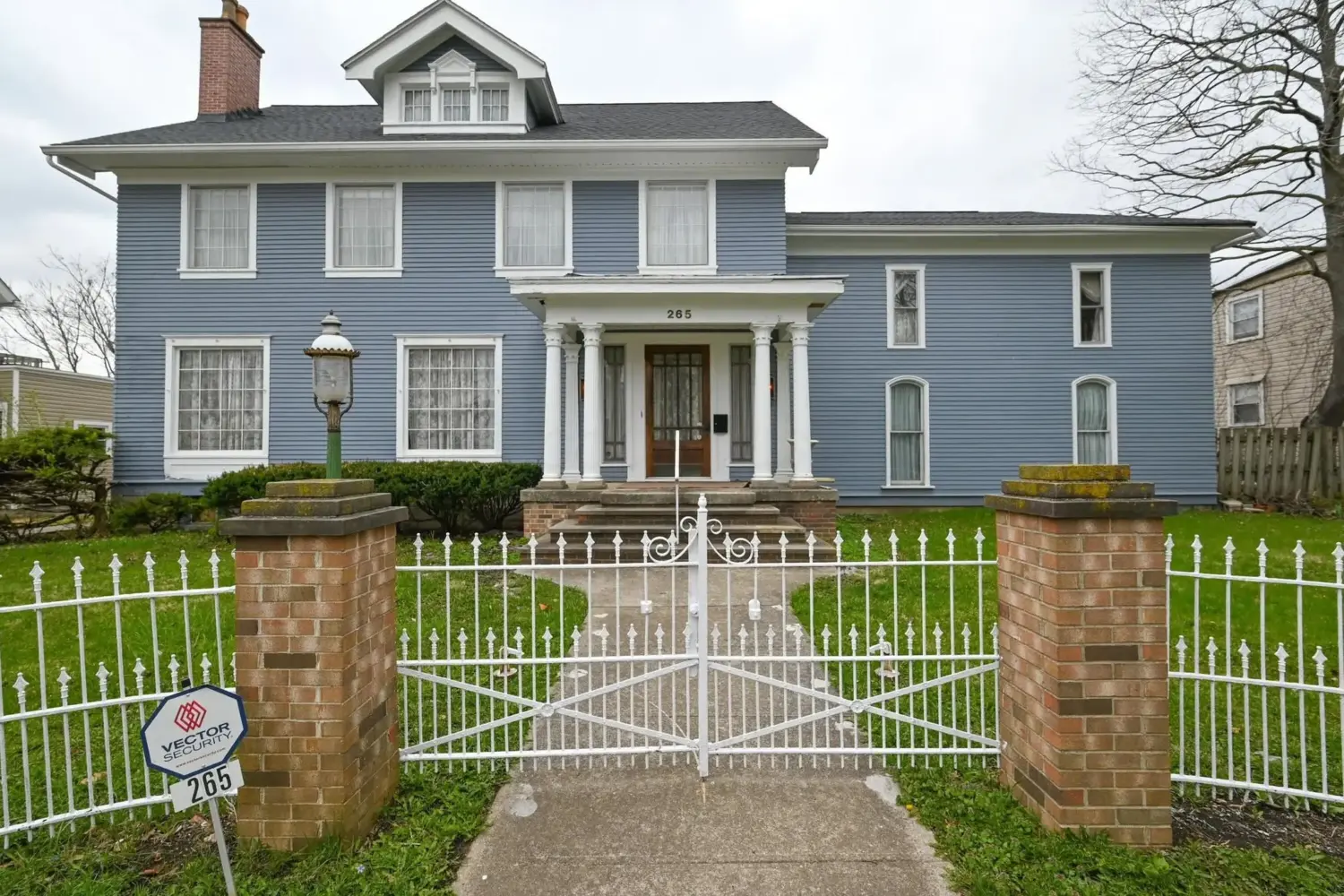 The Olde Towne House is a blue, two-story historic-style building with white trim, tall windows, a central entrance framed by white columns, and a small dormer window above the main roofline. The address, 265, is displayed above the front door. A concrete walkway leads from the sidewalk through a decorative white metal gate between two brick pillars to the front steps. A white metal fence encloses the green lawn, and a vintage-style lamp stands near the entrance. A security sign is posted beside the gate.
