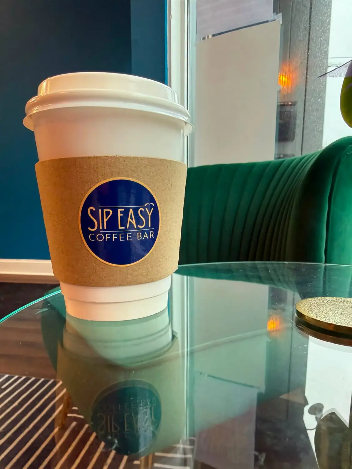 A white takeaway coffee cup with a brown sleeve labeled “Sip Easy Coffee Bar” sits on a round glass table. A dark green upholstered chair and a blue wall are visible in the background, with the cup’s reflection appearing on the tabletop.