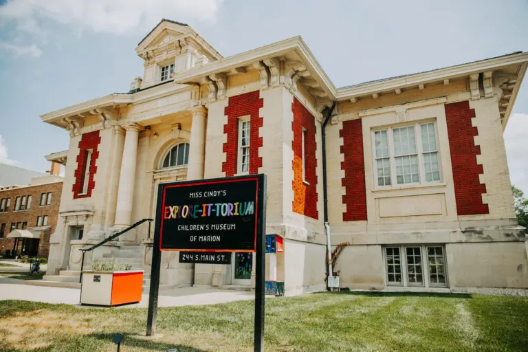 The historic Carnegie Library building in downtown Marion, Ohio, houses the Explore-It-Torium Children’s Museum. The two-story cream-colored brick building features red brick accents, tall windows, decorative columns framing the main entrance, and an ornate tower above the roofline. A black sign with colorful lettering stands on the front lawn, identifying the children’s museum and its location at 244 S. Main St.
