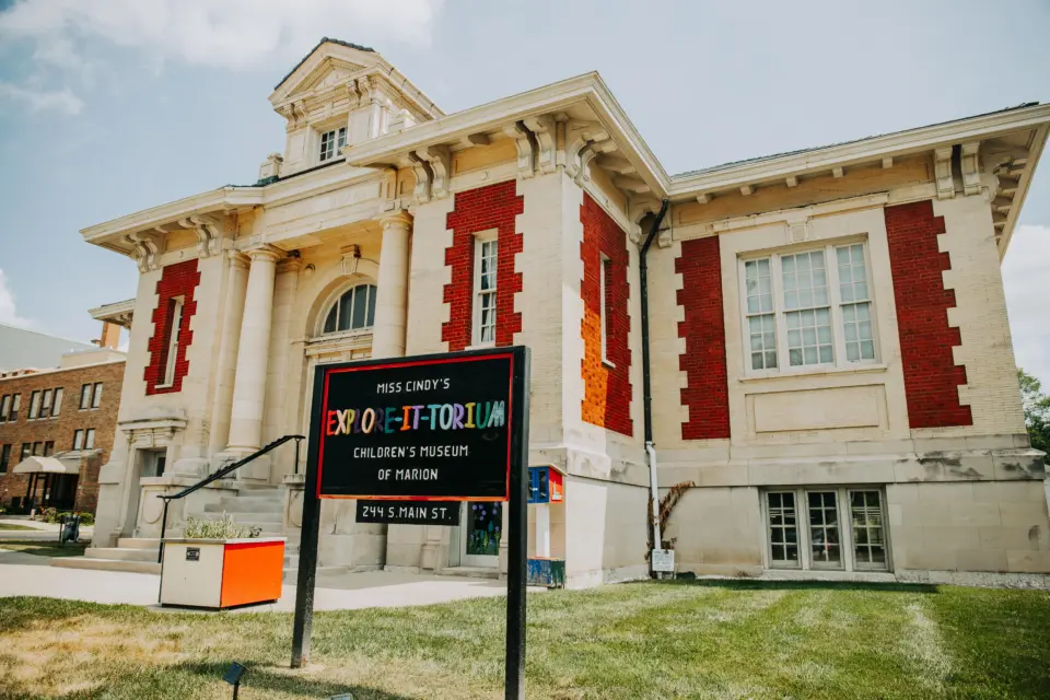 The historic Carnegie Library building in downtown Marion, Ohio, houses the Explore-It-Torium Children’s Museum. The two-story cream-colored brick building features red brick accents, tall windows, decorative columns framing the main entrance, and an ornate tower above the roofline. A black sign with colorful lettering stands on the front lawn, identifying the children’s museum and its location at 244 S. Main St.